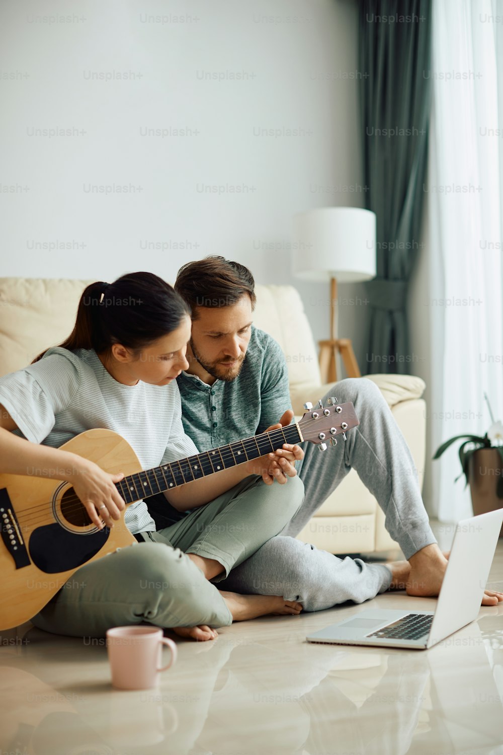 Uomo adulto medio che insegna a sua moglie a suonare la chitarra acustica mentre usa il laptop a casa.