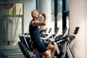 Mature male athlete drinking water after cycling on exercise bike during sports training in a gym.
