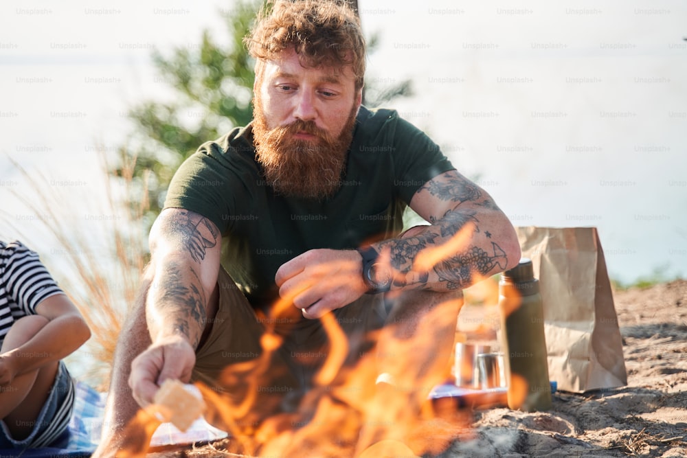 Handsome red haired man frying marshmallow on the flame over the campfire near his daughter. Picnic at the forest. Camping concept