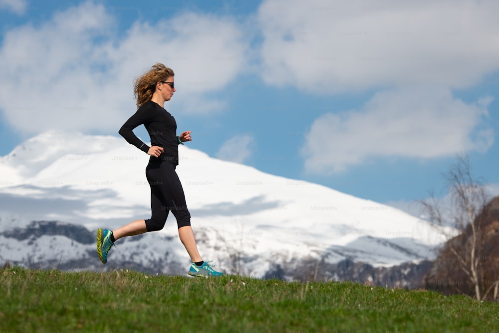 Girl athlete runs in mountains on spring meadow with snow mountains background.