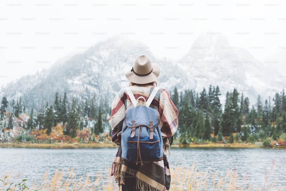 素晴らしい山々の中で幸せを感じるスティッシュな女性は、自然のランドサペをお楽しみください。森と湖、バックパック、帽子、ポンチョを身に着けて、自由奔放に生きるスタイル