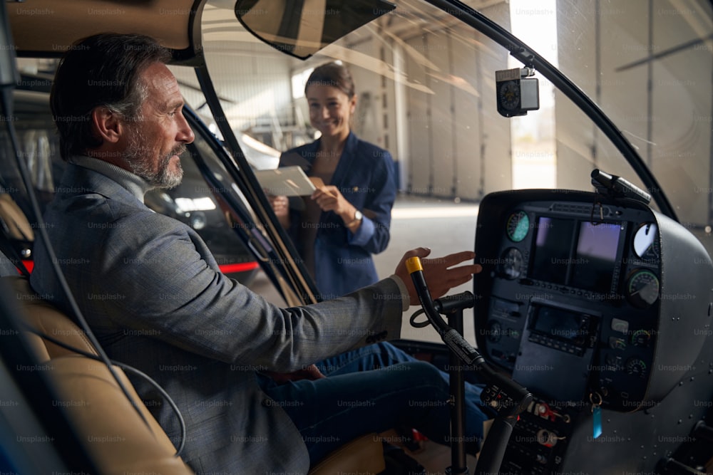Focused client seated in chopper reaching for instrument panel in presence of aircraft hangar manager