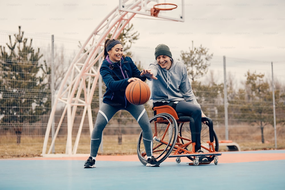 Happy athlete in wheelchair and his female friend having fun while playing basketball outdoors.