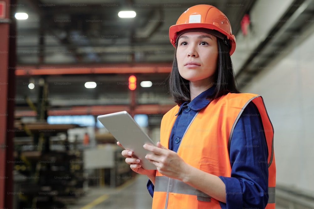 Ingeniera de procesos asiática pensativa con casco de trabajo y chaleco de seguridad usando tableta digital en fábrica