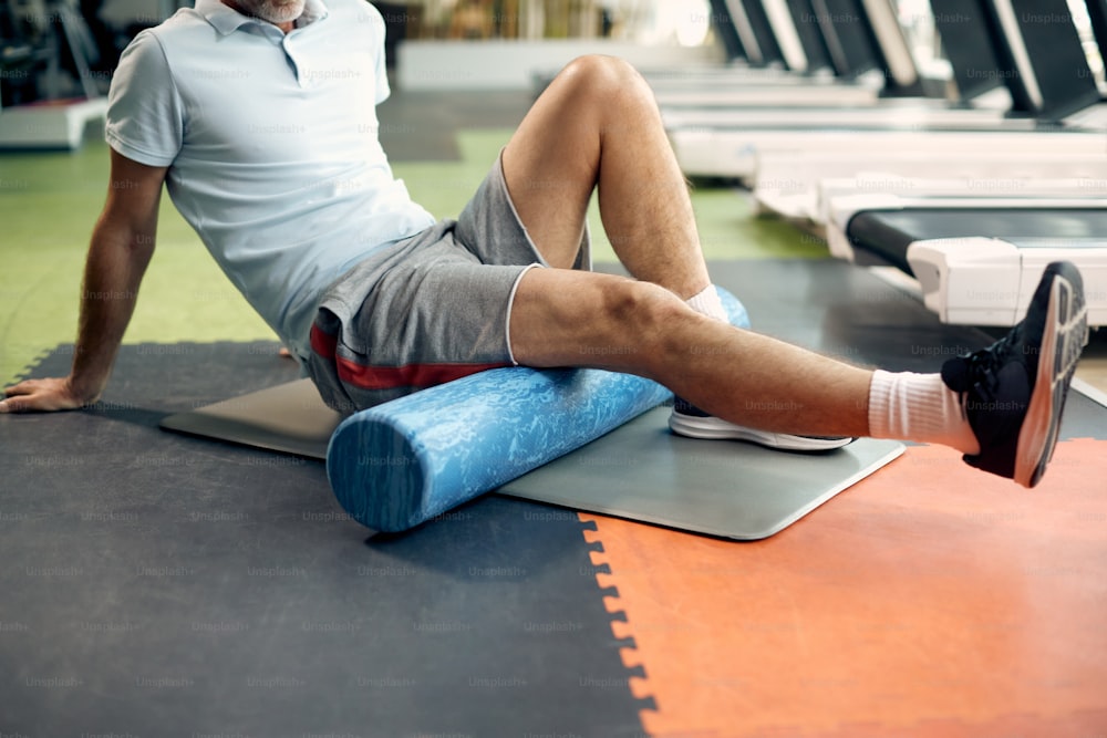 Atleta maduro irreconocible masajeando sus piernas con un rodillo de espuma después de un entrenamiento deportivo en un gimnasio.