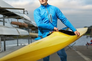 Male athlete in swimming sportswear carrying yellow kayak and paddle outdoors after training on the river, his eyes unseen