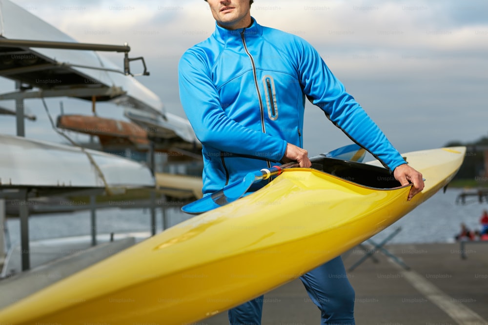 Atleta masculino em roupas esportivas de natação carregando caiaque amarelo e remo ao ar livre após o treinamento no rio, seus olhos invisíveis
