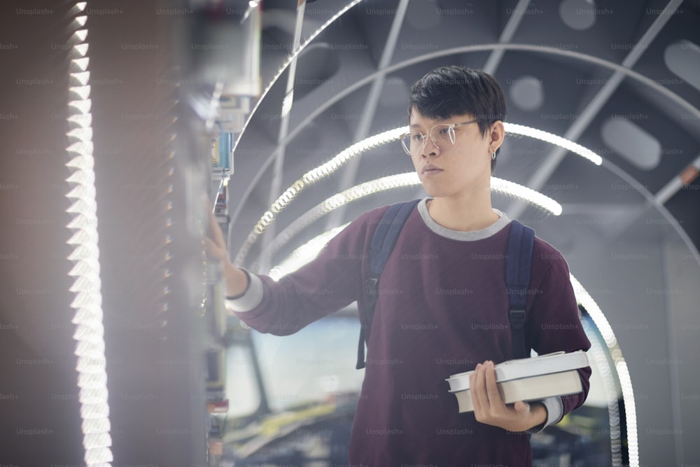 Asian young man in eyeglasses standing near the bookshelf and choosing books for reading in the library