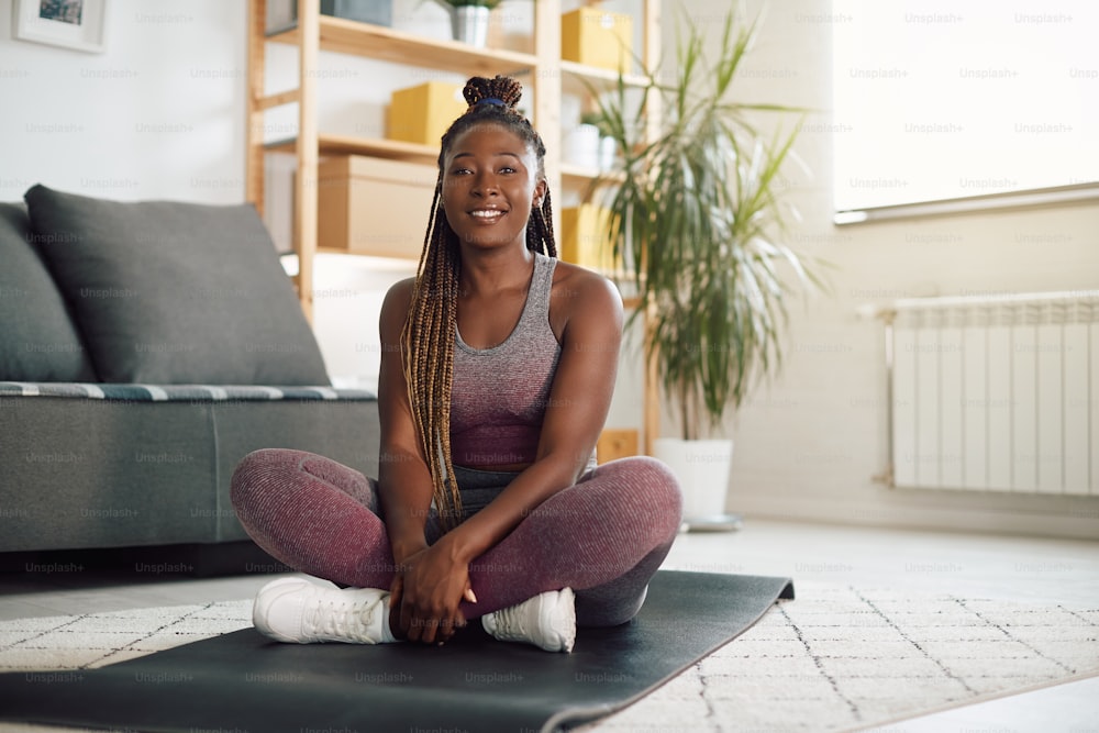 Portrait of black female athlete sitting on exercise mat in the living room and looking at camera.