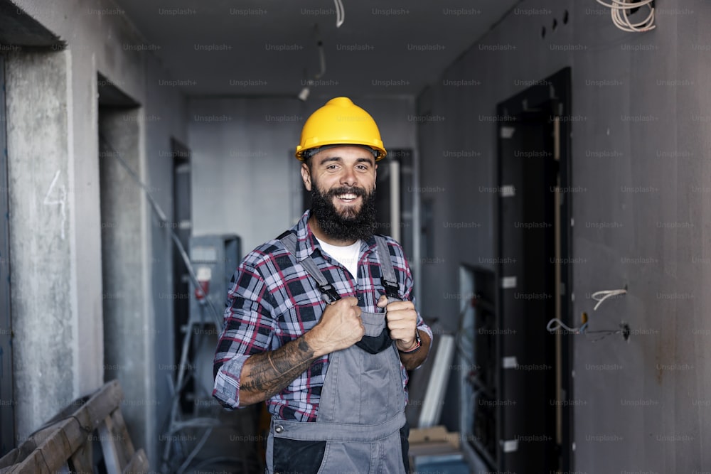 A happy, hardworking handyman with a helmet on his head holds his overalls and looking at the camera. There is a lot of work to do, but he is inspired and happy to get to work.