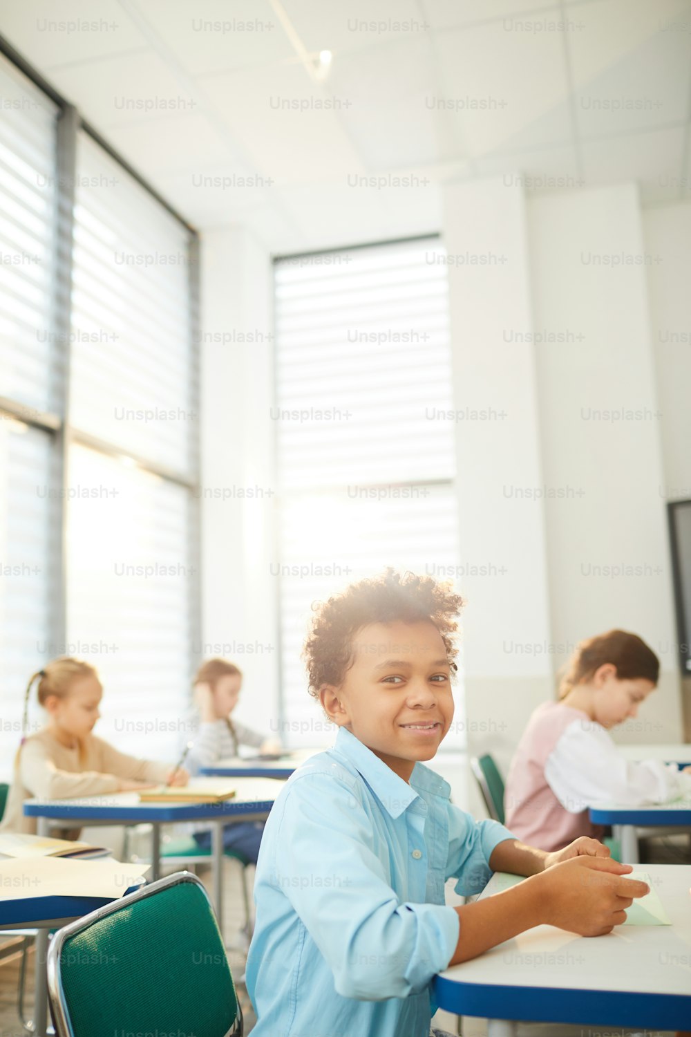 Ritratto verticale di allegro ragazzo di razza mista con capelli crespi seduto al banco di scuola che guarda la macchina fotografica sorridente, spazio di copia