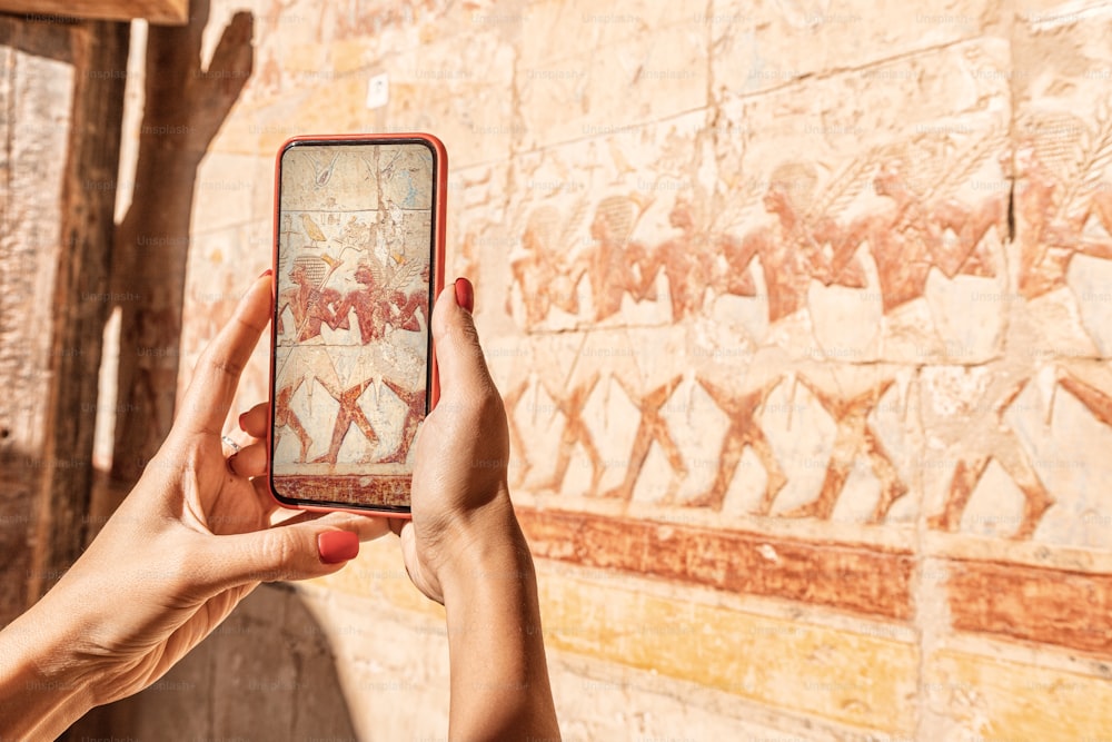 여행 블로거 소녀는 이집트의 고대 도시 룩소르에있는 유명한 하트셉수트 사원 프레스코 화에서 스마트 폰으로 사진을 찍습니다. 또는 시각적 관광 가이드 사용