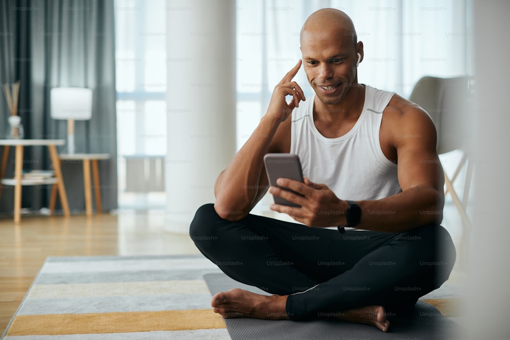 Feliz atleta afroamericano usando el teléfono celular y escuchando música mientras se relaja después de hacer ejercicio en casa.