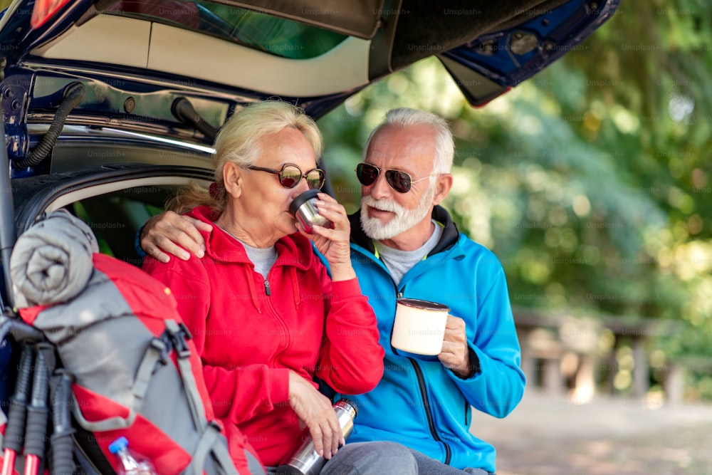 Senior couple sitting in the trunk and drink coffee while resting from hiking.