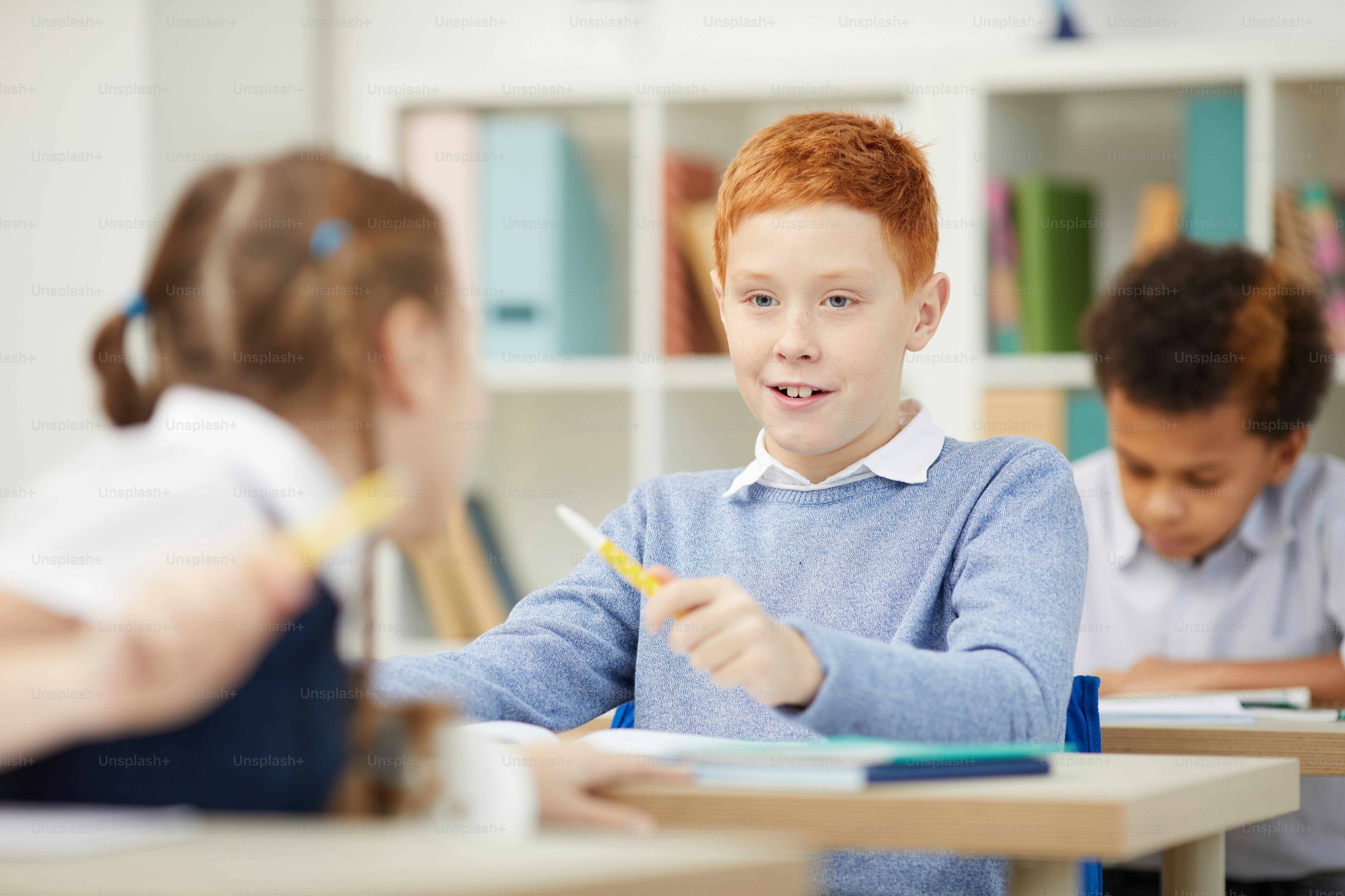 Red haired schoolboy talking to his classmate while they sitting at desks during a lesson in the classroom