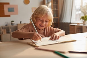 Niño pequeño sentado a la mesa y tomando notas en el cuaderno haciendo su tarea en casa