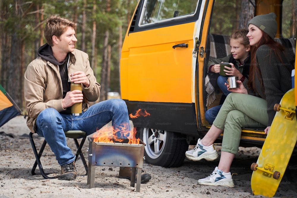 Smiling father opens a thermos with tea and squints against the bright sun. Happy family sitting at the nature and drinking hot tea from cups at camp fire in cozy forest