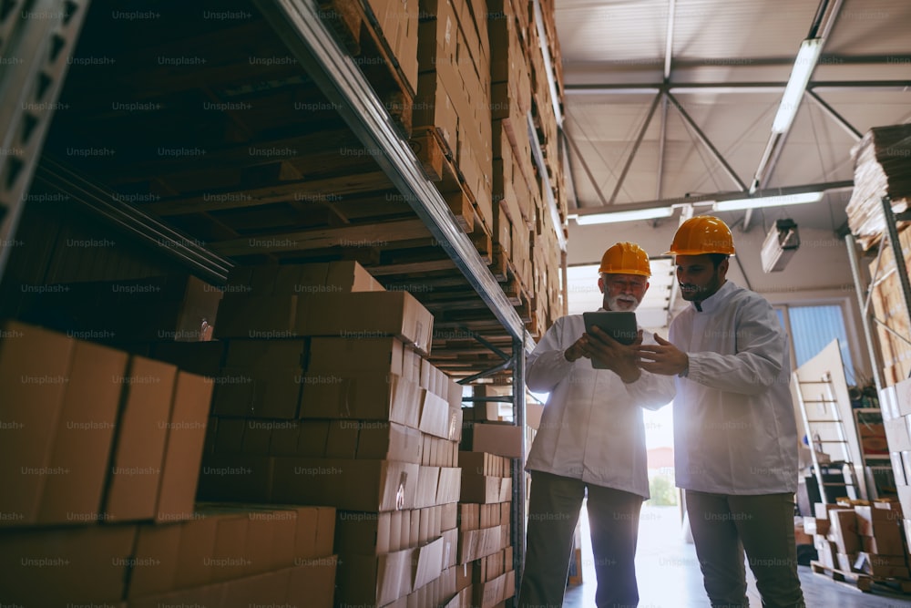 Two hardworking warehouse workers standing in warehouse and checking inventory. Older one holding tablet while younger one looking at it.