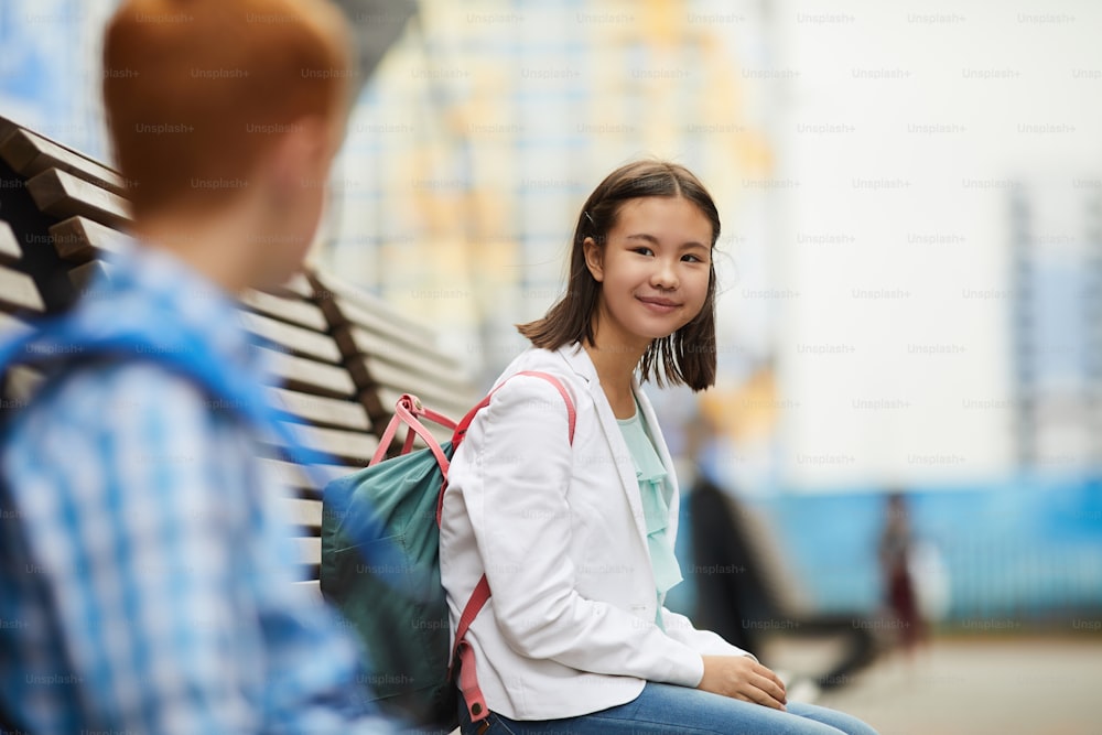 Asian schoolgirl with backpack sitting on the bench and talking to her classmate after school outdoors