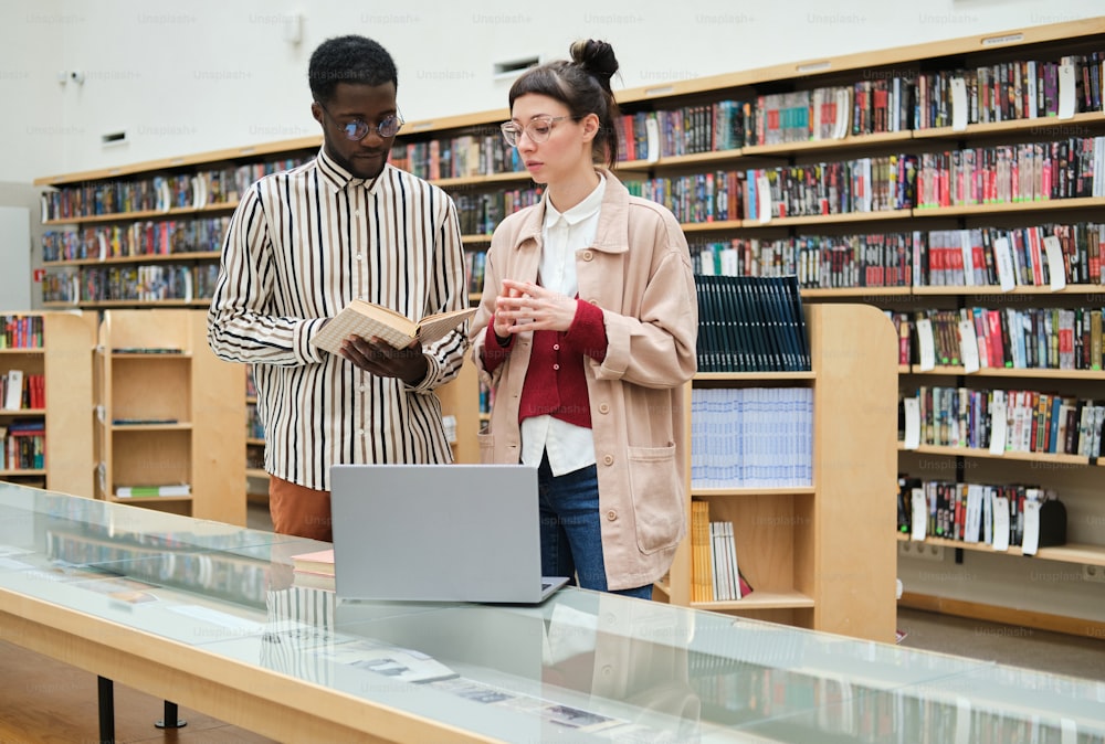 African young man consulting with woman and choosing a book for reading in the library