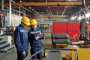 Two male engineers in workwear standing in the middle of large workshop or plant and discussing characteristics of new industrial equipment