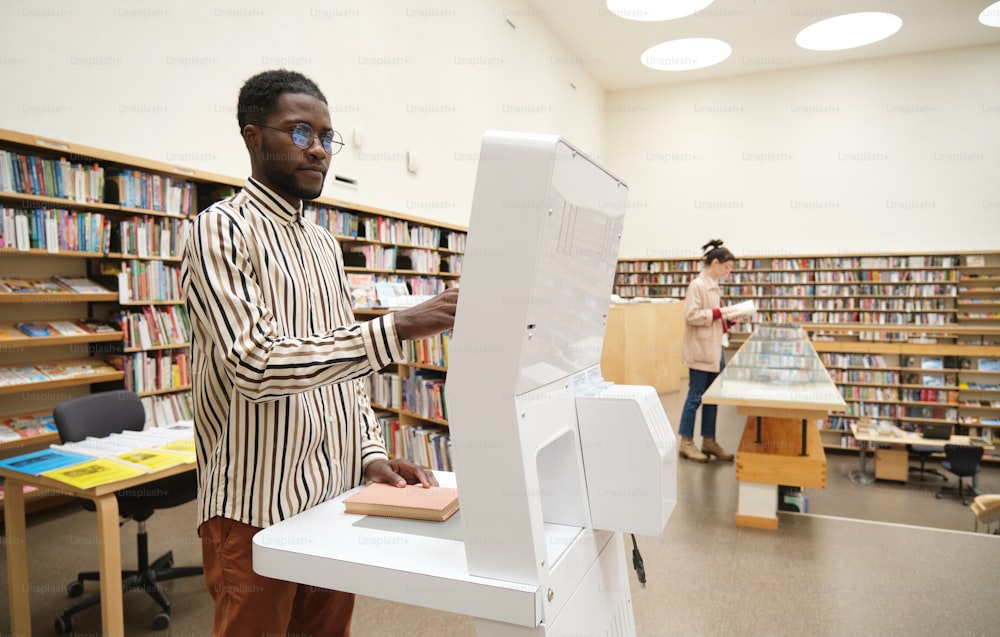 Joven africano de pie en la biblioteca y eligiendo libros para leer