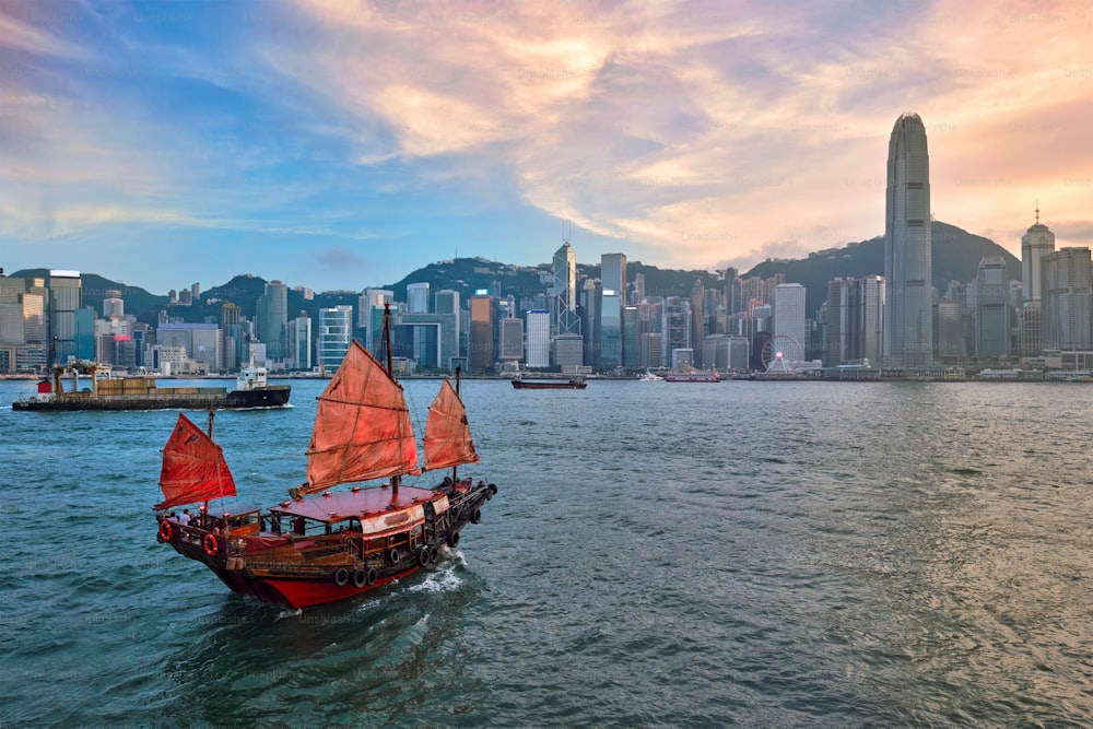香港のスカイライン、街並み、ダウンタウンの高層ビル、夕方にはビクトリアハーバー、夕暮れ時のジャンク観光フェリーボート、ドラマチックな空。香港、中国