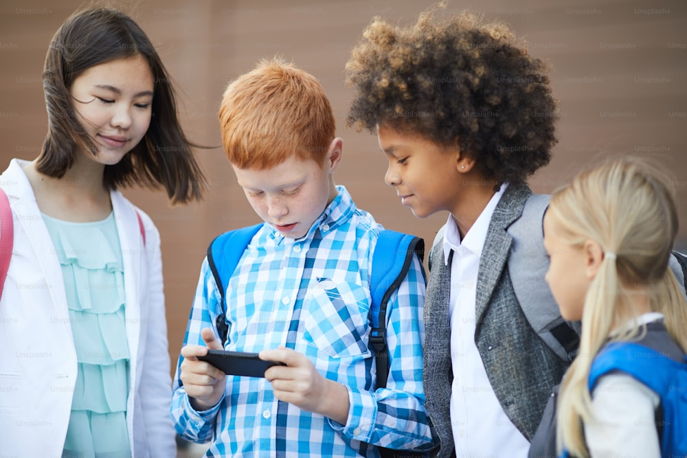 Schoolboy with backpack playing game on his mobile phone while standing together with his classmates after lessons at school