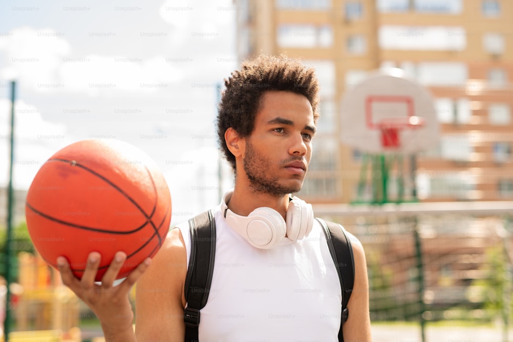 Serious active guy with ball standing on basketball court or playground during outdoor training