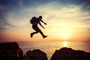 Tapferer Mann mit Rucksack, der bei Sonnenuntergang über Felsen in der Nähe des Ozeans springt
