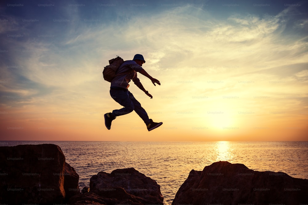 brave man with backpack jumping over rocks near ocean in sunset