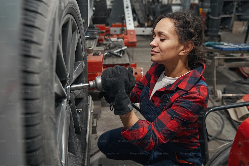 Female vehicle technician in work gloves fixing wheel in automobile repair garage