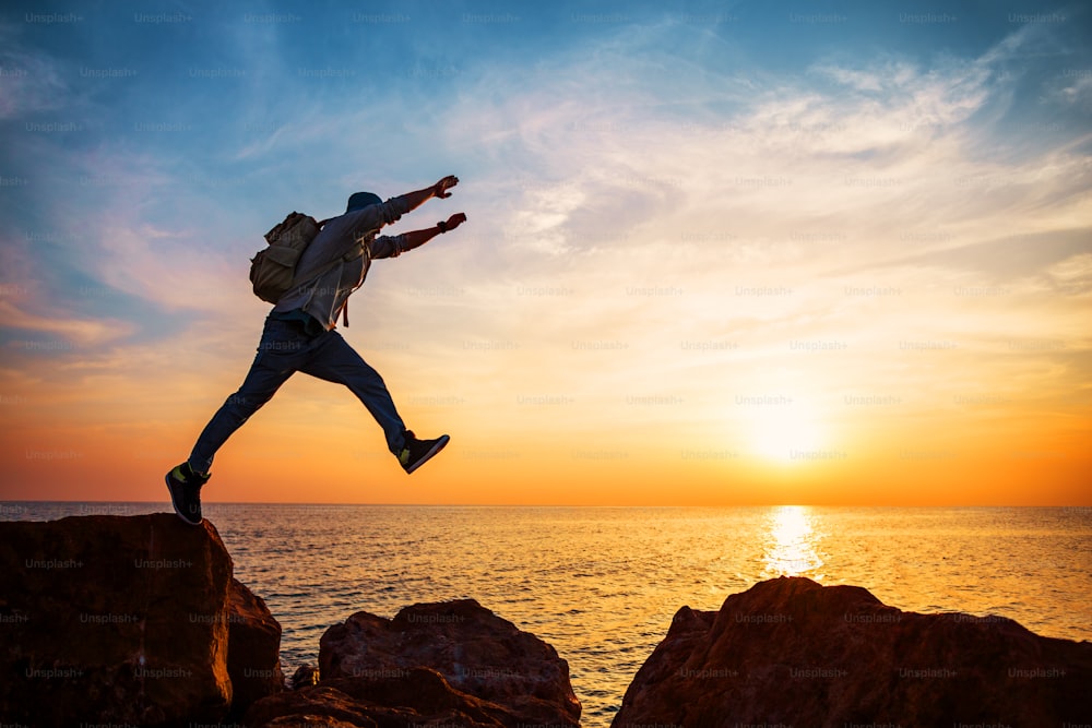 homem corajoso com mochila saltando sobre rochas perto do oceano no pôr do sol