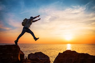 brave man with backpack jumping over rocks near ocean in sunset