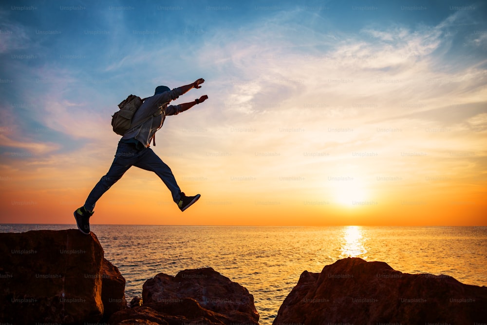 brave man with backpack jumping over rocks near ocean in sunset