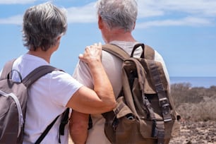 Back view of couple of senior travelers in outdoors excursion in arid landscape wearing backpack looking at horizon over sea
