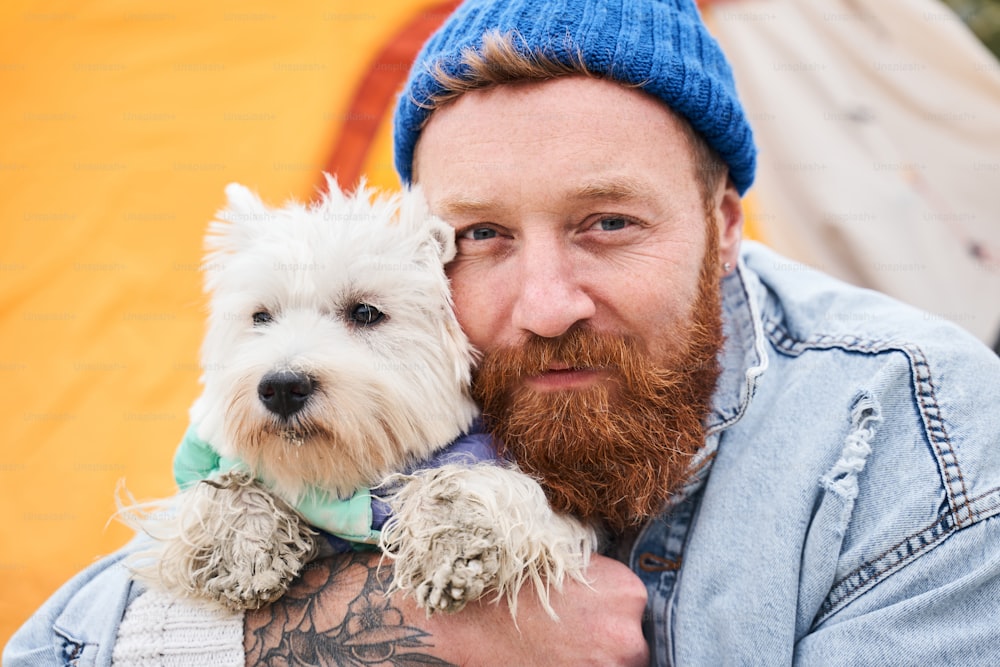 Look at us! Bearded man looking to the camera while cuddling his white terrier dog. They are best friends. Stock photo