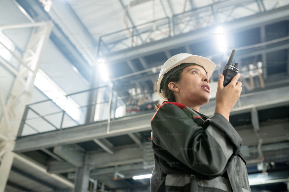 Young female engineer in protective helmet and workwear using walkie talkie while standing in large workshop of contemporary factory
