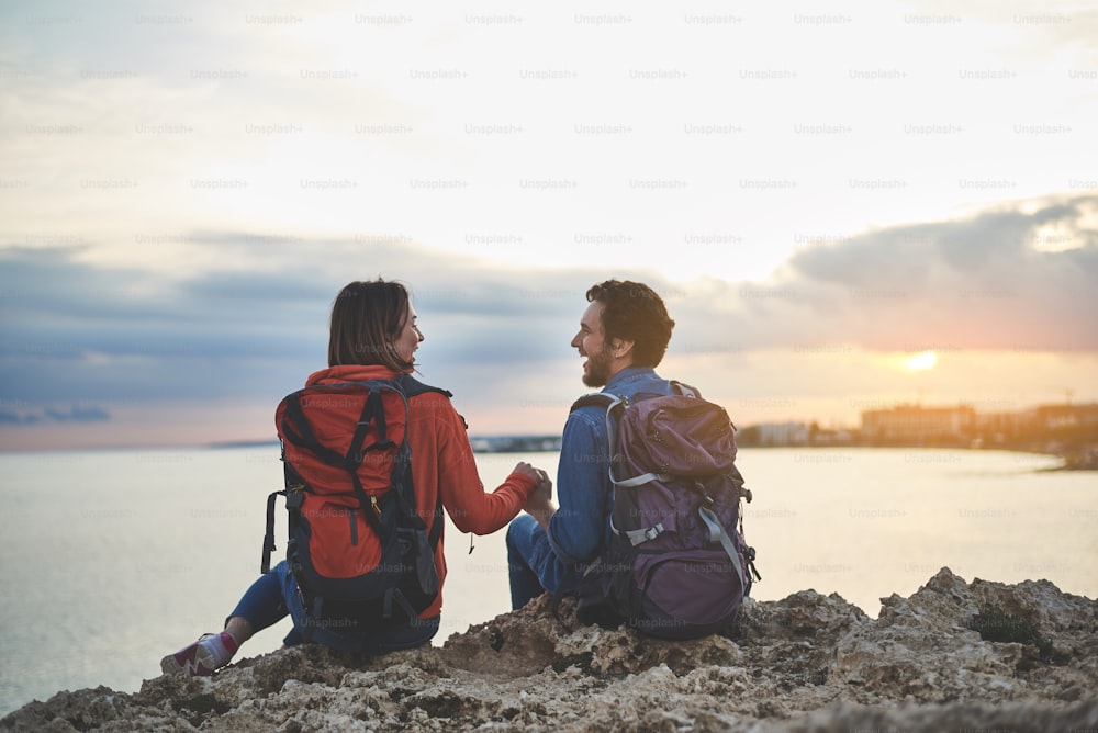 Una feliz pareja joven y amorosa está viendo la puesta de sol cerca del océano. Están sentados en la roca y riendo mientras se toman de la mano