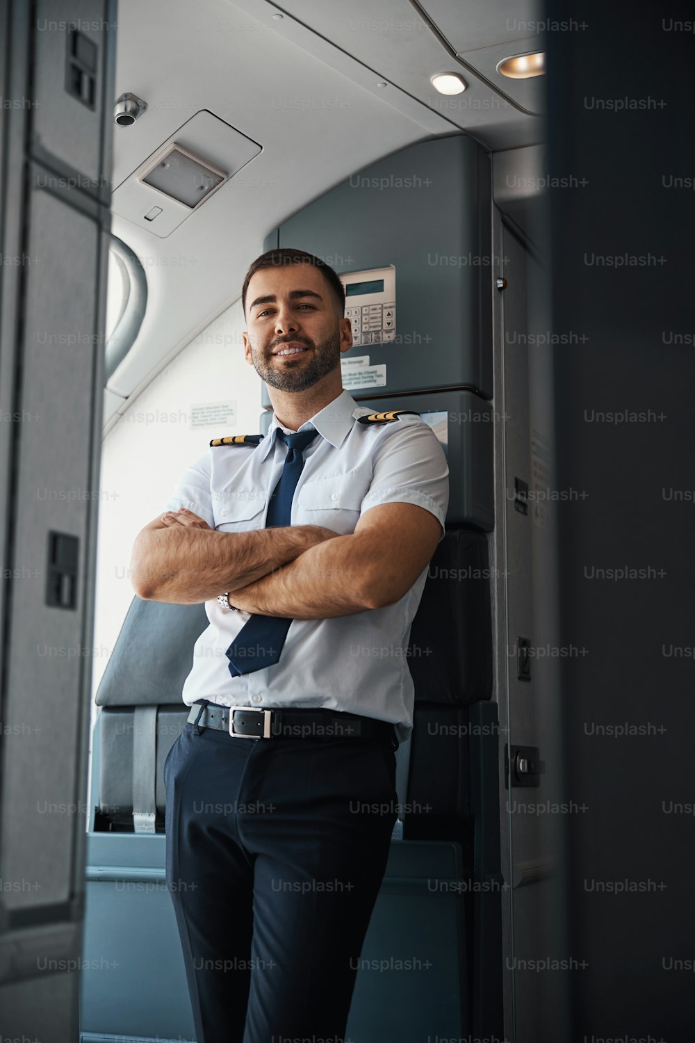 Cheerful Caucasian pilot having his arms crossed while standing by the wall in an airplane and smiling