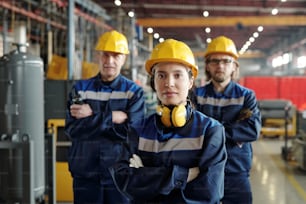 Portrait of content young female plant worker with sound-proof headphones standing with crossed arms against colleagues in compressor factory shop