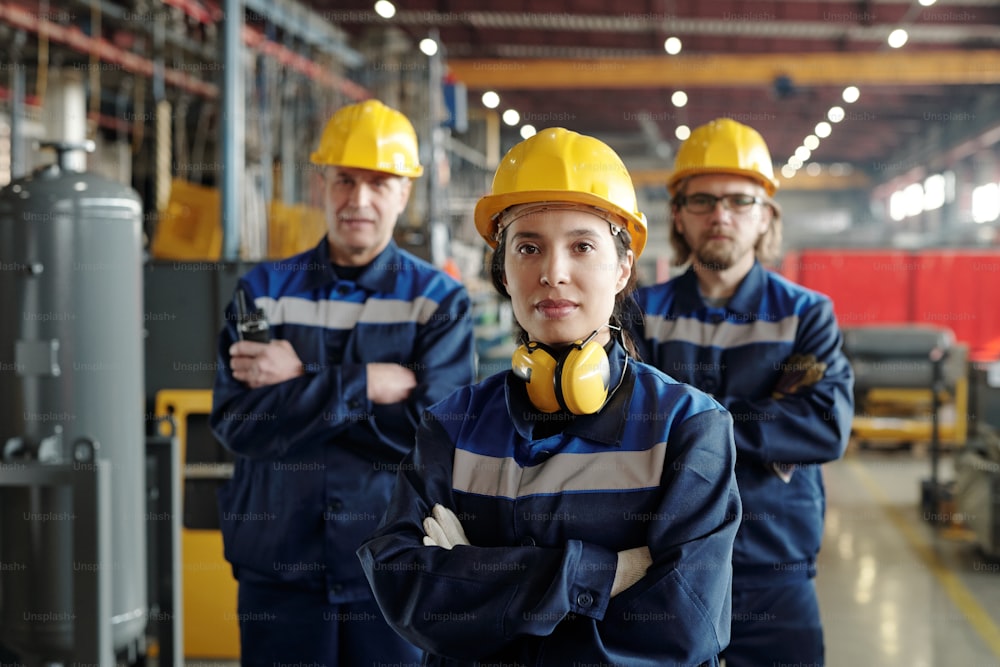 Retrato do conteúdo da jovem trabalhadora da fábrica com fones de ouvido à prova de som em pé com os braços cruzados contra colegas na loja da fábrica de compressores