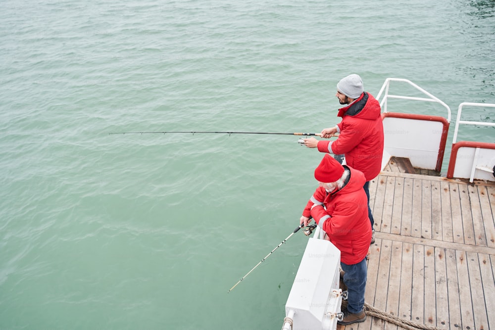 Vista de alto ângulo do jovem marinheiro ajudando o pescador diligente a tirar peixes enormes do mar ou do oceano durante a pesca no dia de inverno. Foto de Stock
