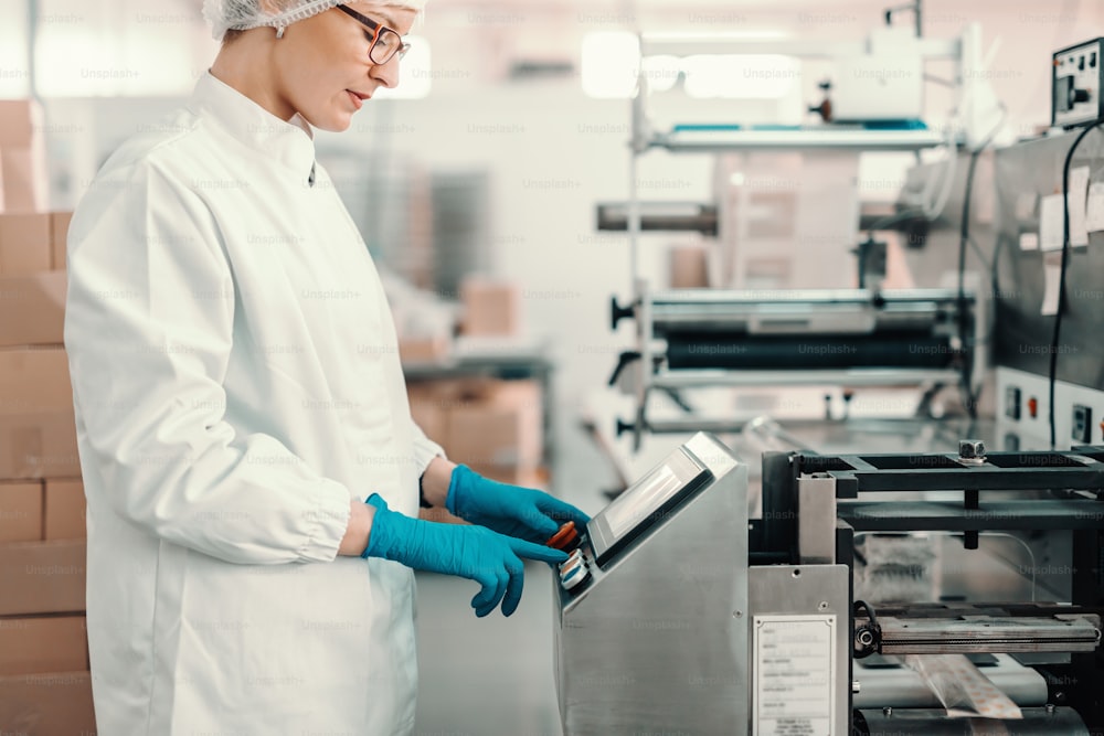 Junge Mitarbeiterin in steriler Uniform und blauen Gummihandschuhen schaltet die Verpackungsmaschine ein, während sie in der Lebensmittelfabrik steht.