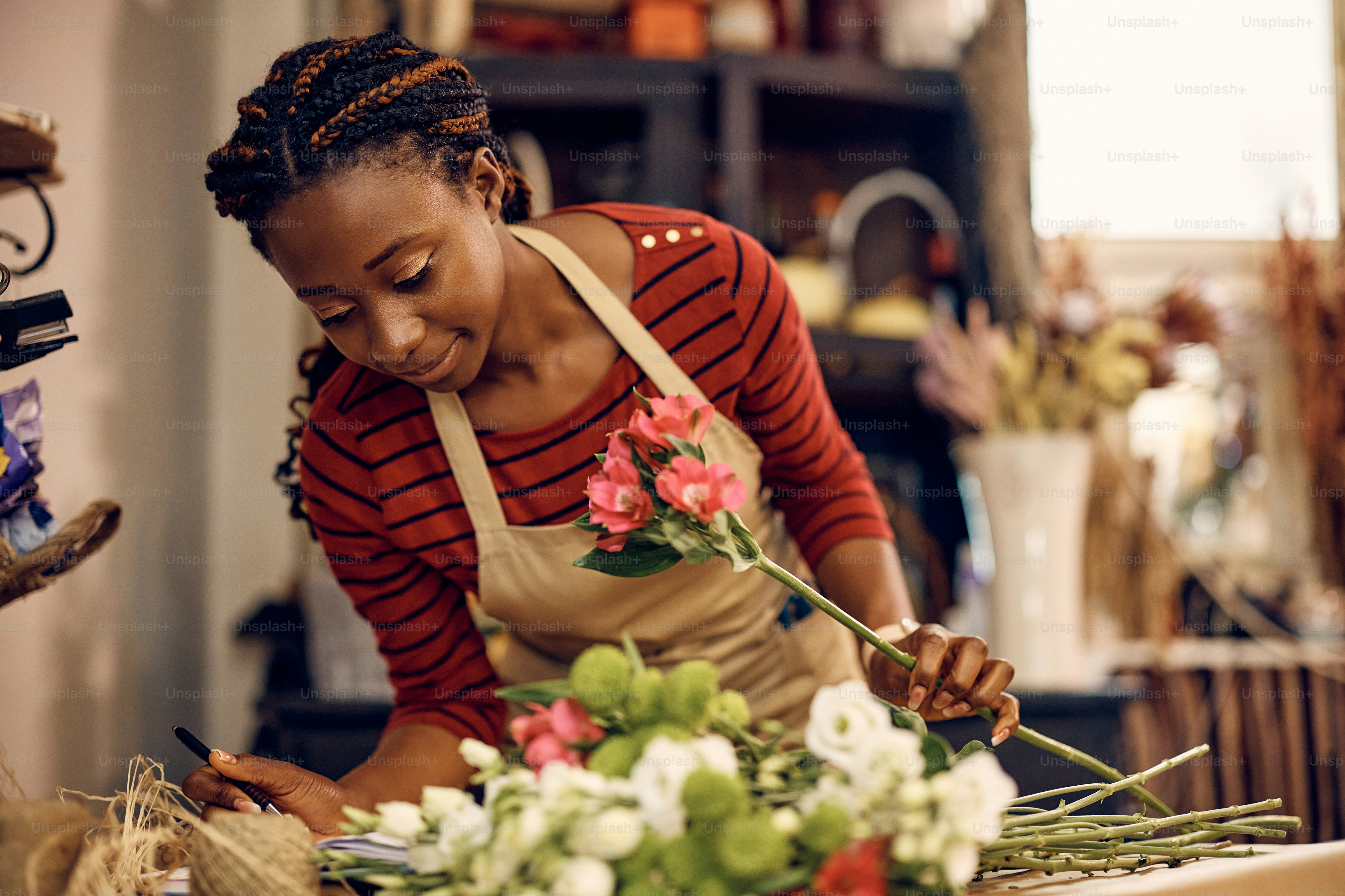 Florista afroamericana leyendo notas de pedido mientras hace un ramo de flores en su floristería.