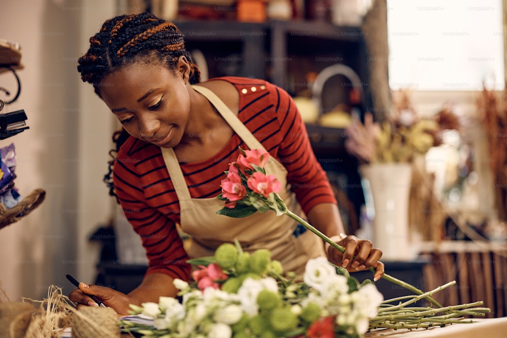 Une fleuriste afro-américaine lit des notes de commande tout en préparant un bouquet dans son magasin de fleurs.