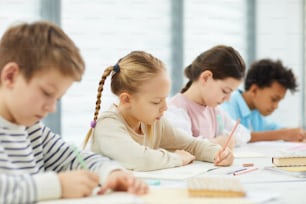 Horizontal portrait of four children wearing casual outfits sitting at desk in modern classroom writing something in their notebooks, copy space