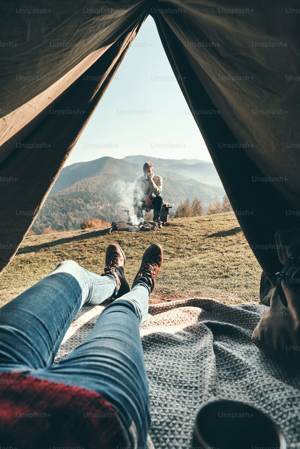 Frau genießt den Blick auf die Bergkette vom Zelt aus, während ihr Freund sich am Lagerfeuer ausruht