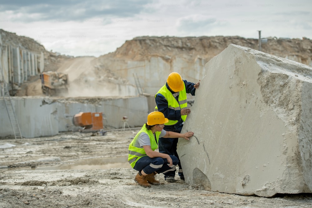 Due giovani muratori in elmetto e uniforme che lavorano vicino a un enorme pezzo di cemento