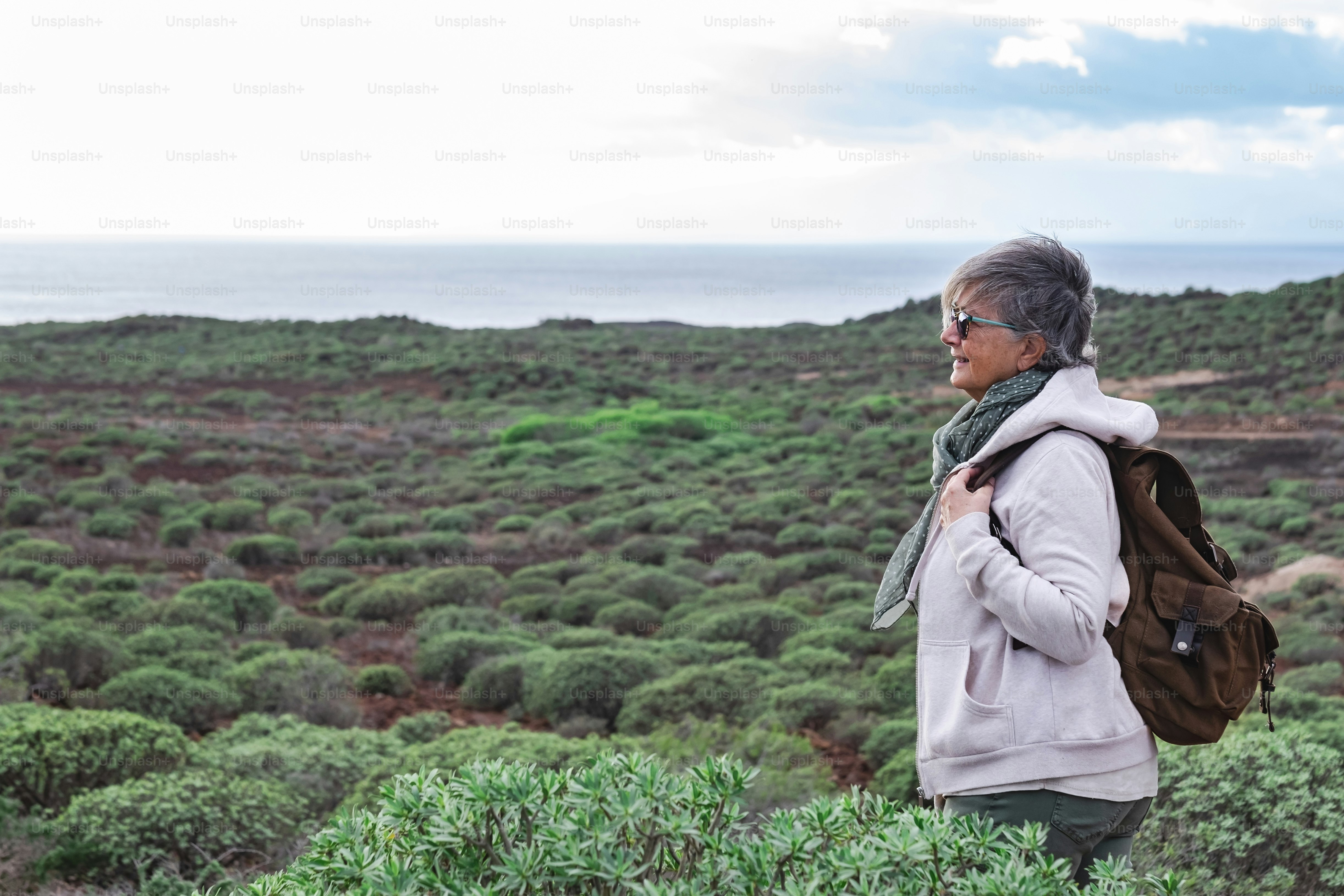 Lächelnde ältere Frau, die einen Ausflug im Freien zwischen grünen Büschen und Meer genießt. Grauhaarige ältere Menschen mit Rucksack mit Blick auf den Horizont